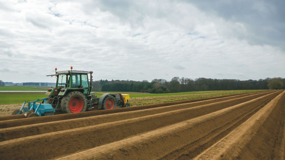 Un tracteur laboure des rangées de terre dans un champ agricole sous un ciel nuageux.